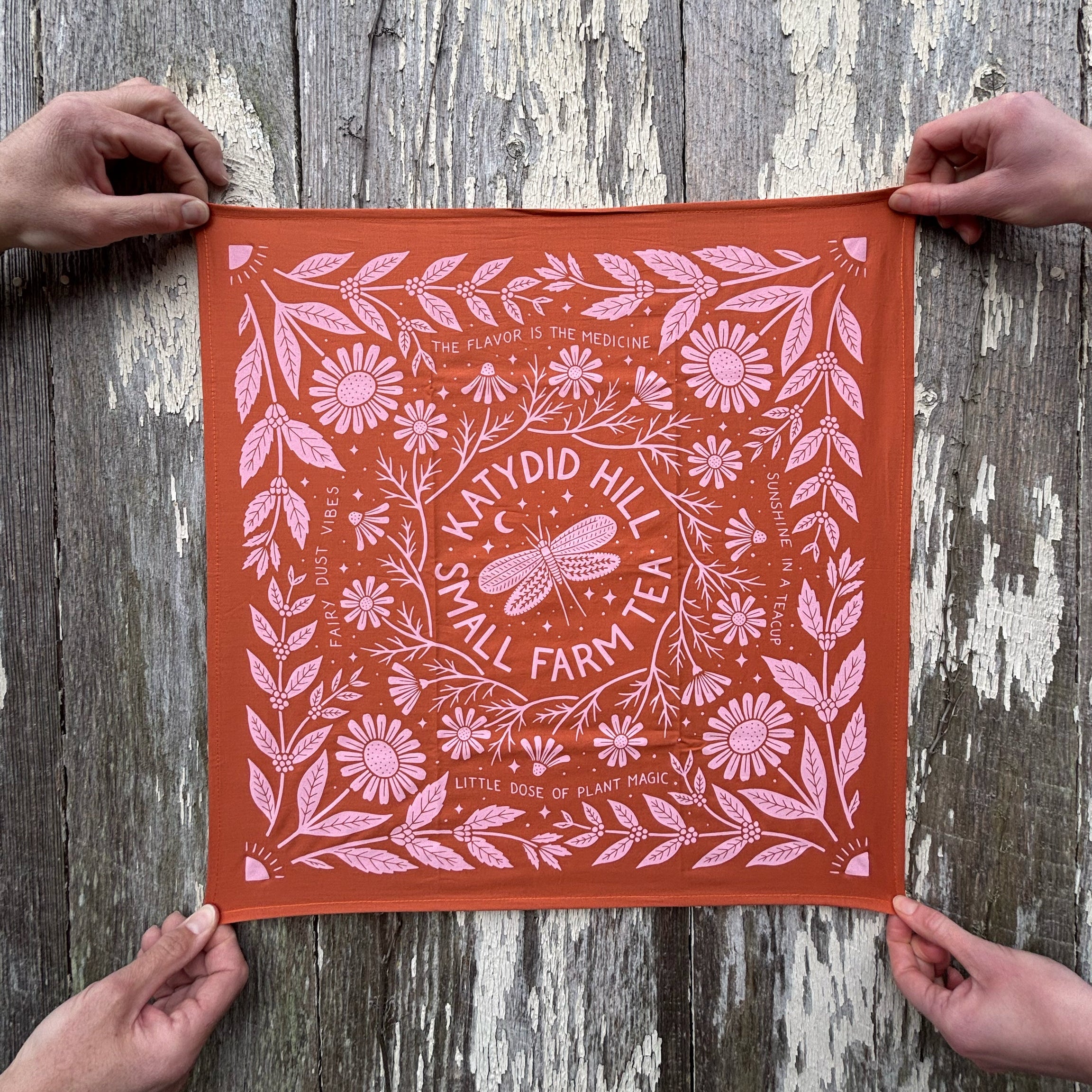 Orange fabric with pink floral and katydid pattern held by two hands against a wooden background