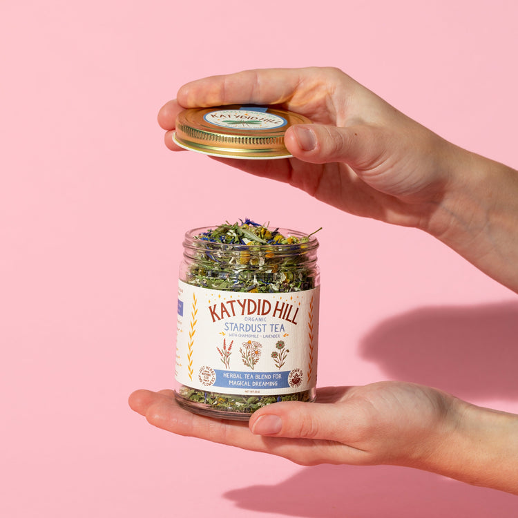 hands holding an open glass jar of stardust loose leaf tea on pink background