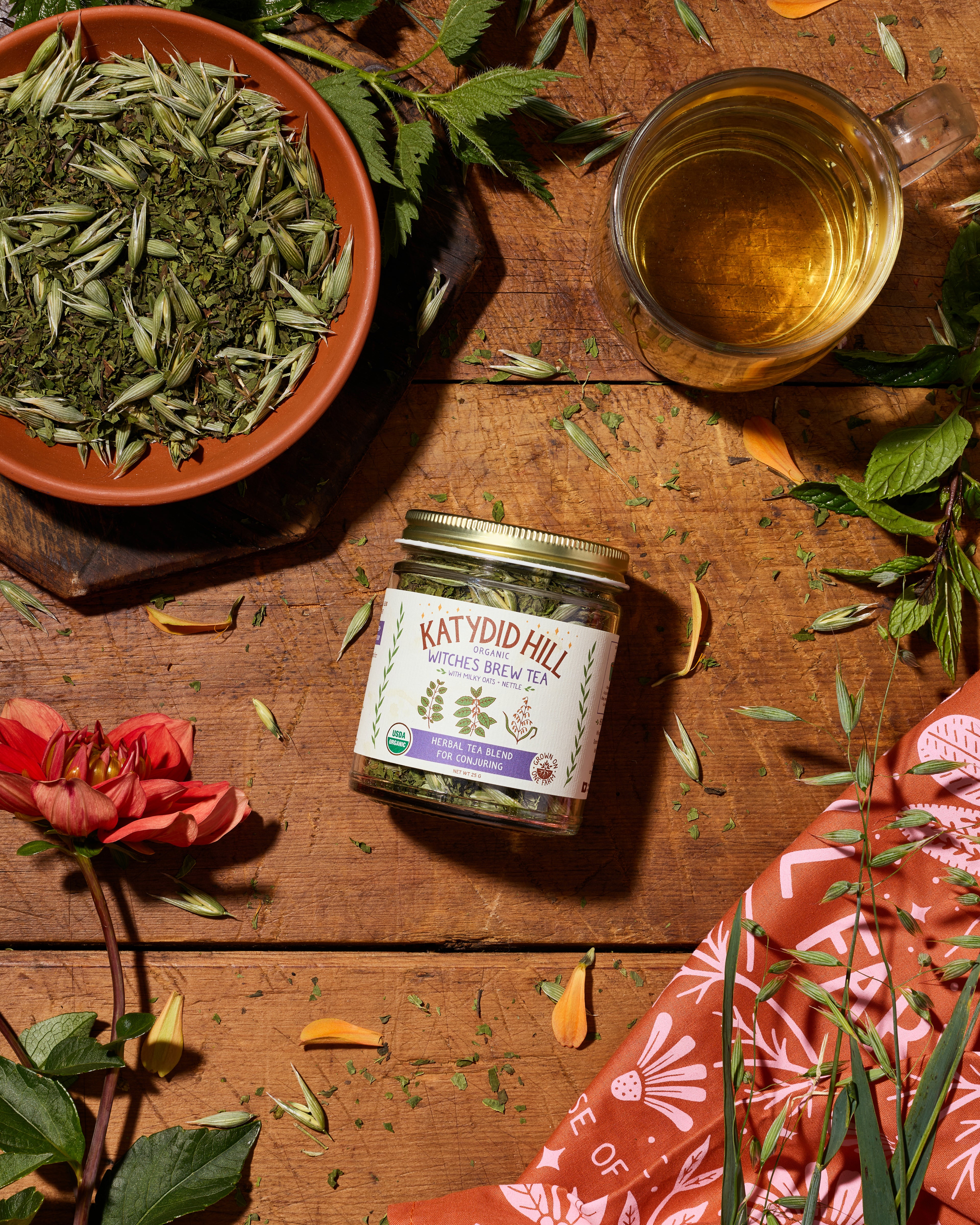 Jar of Witches Brew tea herbal tea blend on a wooden surface with a cup of tea and a bowl of milky oats. Herbal Tea blend for conjuring with milky oats and nettle.