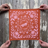 Orange fabric with pink floral and katydid pattern held by two hands against a wooden background