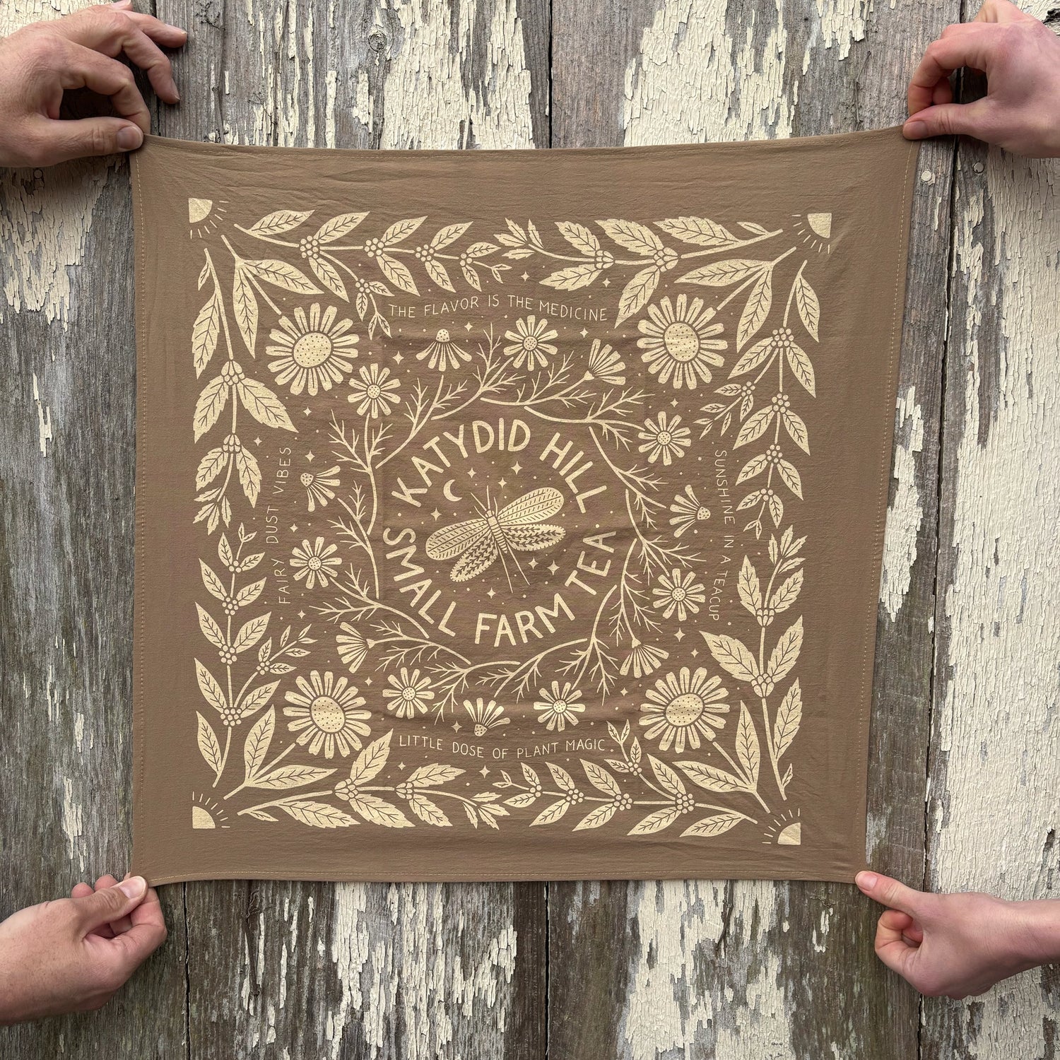 Brown fabric with floral and katydid pattern held by two hands on a wooden surface