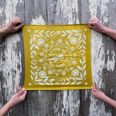 Yellow fabric with white floral pattern held by four hands against a wooden background