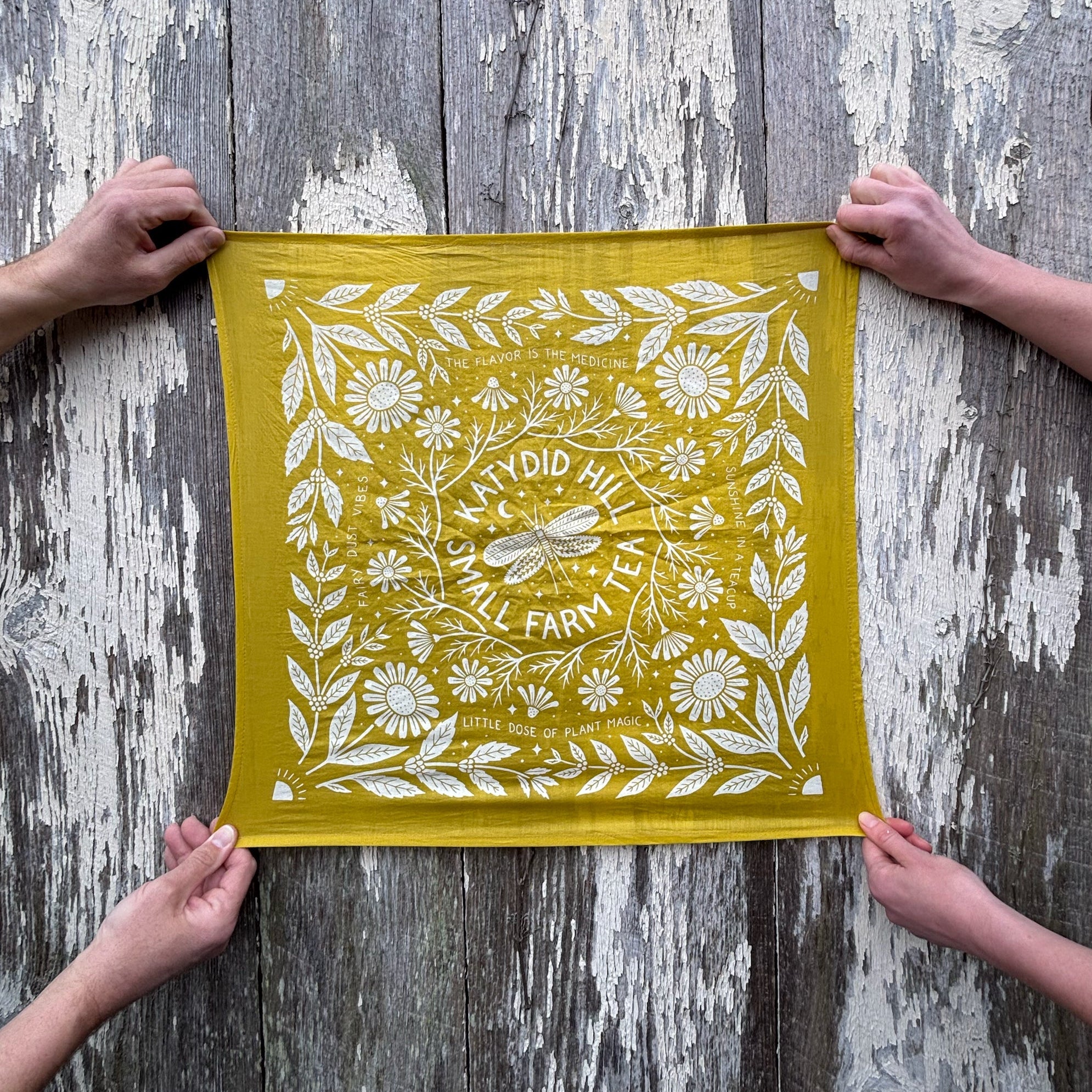 Yellow fabric with white floral pattern held by four hands against a wooden background